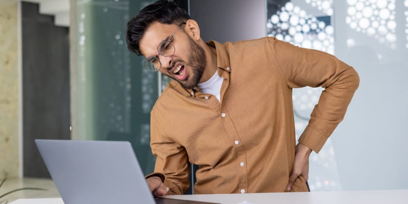 A young man working on his laptop experiencing severe lower back pain.