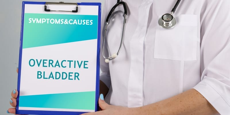Doctor holding a notepad displaying the text 'Overactive Bladder Symptoms & Causes'.